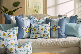 A couch adorned with pastel blue and white pillows featuring bee designs is complemented by the Blossoming Peonies Lumbar Pillow from the Bee & Florals Collection, showcasing yellow flowers and green leaves. A plant and framed picture adorn the background, enhanced by natural light.