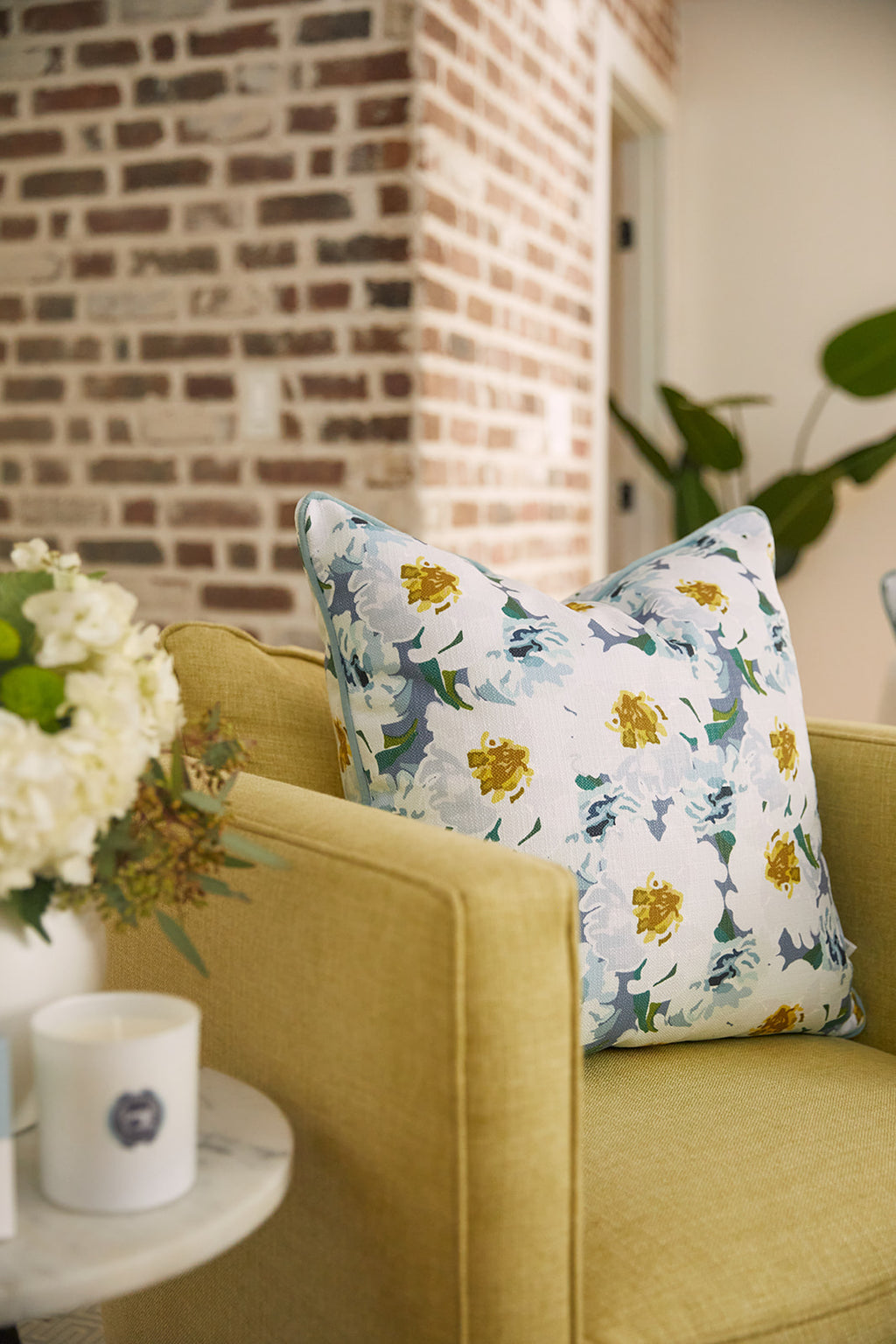 A cozy living space with a yellow armchair featuring the Bee & Florals Collection: Blossoming Peonies Pillow from Sewing Down South®, showcasing blue and yellow flowers. A small side table holds a white candle and floral arrangement, while a rustic brick wall adds warmth to the setting.