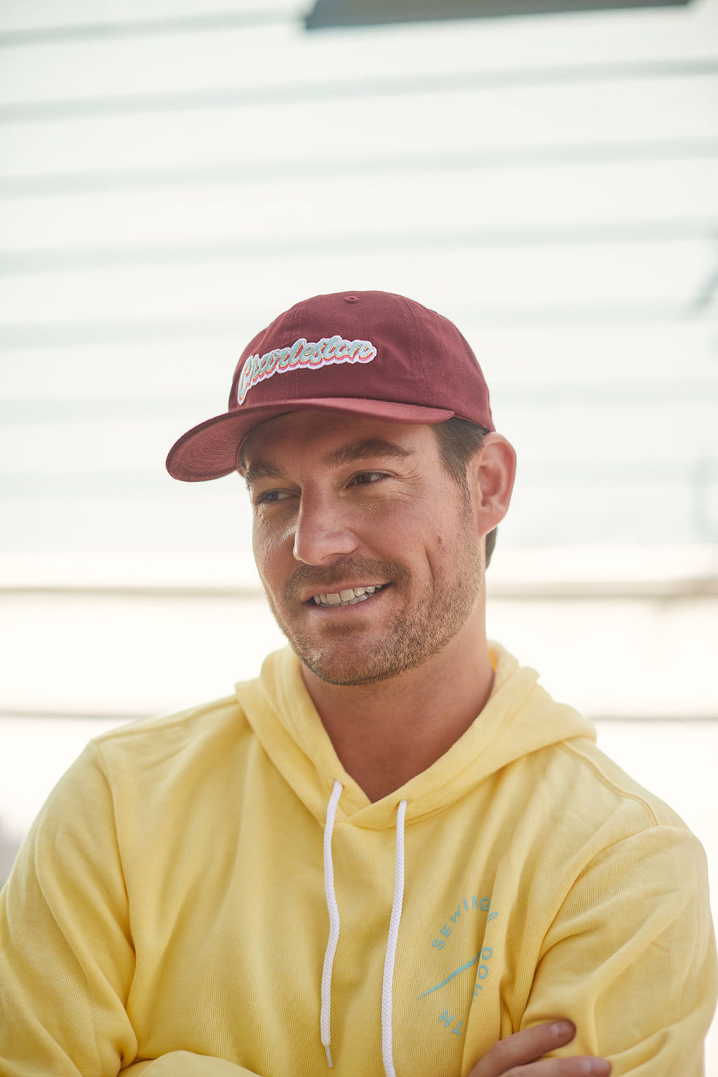 A man in a yellow hoodie and a Charleston Dad Cap in Maroon with an adjustable backstrap smiles outdoors. The background features a light-colored building with horizontal siding, highlighting the premium cotton of his stylish headwear.