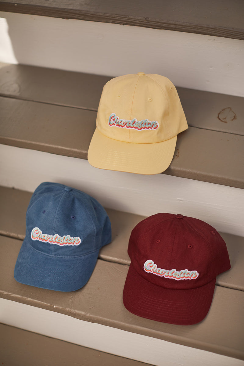 Three Charleston Dad Caps in premium cotton are displayed on the steps: a yellow cap on top, a blue one in the middle, and a red one at the bottom. Each features Chevalier embroidered in colorful letters and includes an adjustable backstrap for a perfect fit.