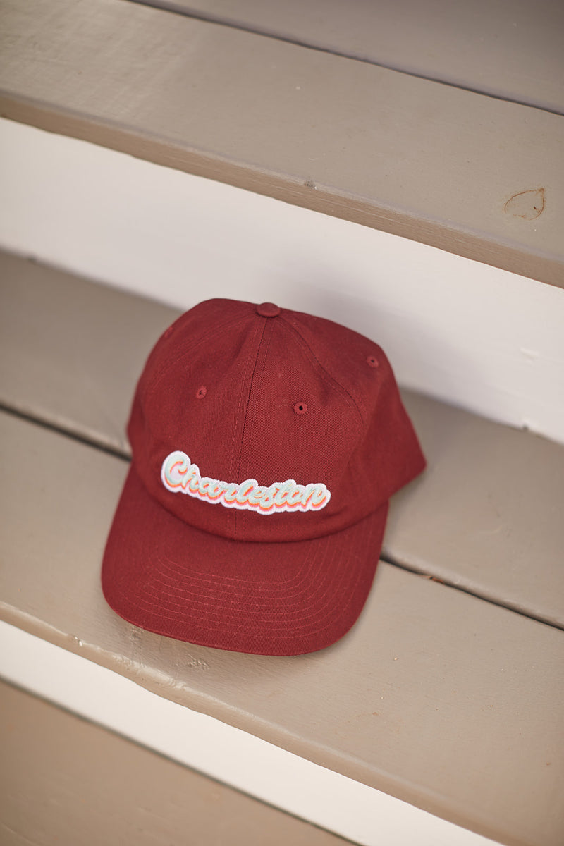 A maroon Charleston Dad Cap, made from premium cotton with Charleston in white script, rests on beige wooden steps. Its adjustable backstrap ensures a comfortable and stylish fit for anyone.