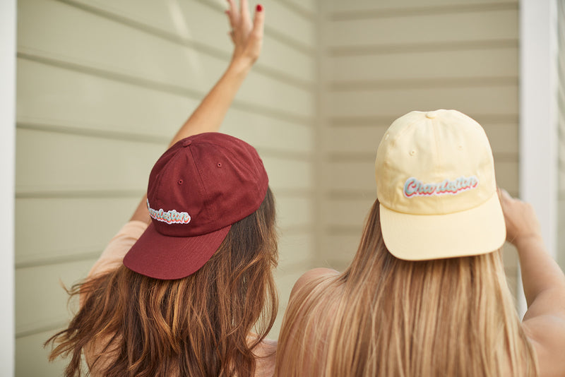 Two individuals with long hair wear light yellow and Charleston Dad Cap in maroon, facing a beige wall. One raises an arm, highlighting the premium cotton texture and adjustable backstrap of their embroidered caps, their faces concealed.