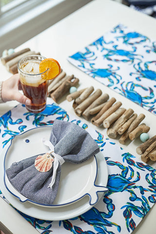 A person holds iced tea with an orange slice at a coastal-inspired table. The scene includes Reversible Crab Craze Placemats and a plate with a seashell napkin, surrounded by driftwood and stones for added serenity.