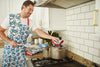 Wearing a patterned apron, a man prepares food in a kitchen. Using the Crab Craze pot holder with its heat-resistant silicone grip, he lifts the lid of a pot on the stove. The kitchen features white cabinets and a sleek tiled backsplash.