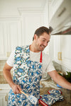 A man in a white t-shirt and crab-patterned apron smiles while cooking in his coastal-themed kitchen. With Sewing Down South’s™ Crab Craze Kitchen Towels over his shoulder, he stands among white cabinets with gold handles, poised for culinary magic.