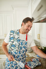 A man in a white t-shirt and crab-patterned apron smiles while cooking in his coastal-themed kitchen. With Sewing Down South’s™ Crab Craze Kitchen Towels over his shoulder, he stands among white cabinets with gold handles, poised for culinary magic.