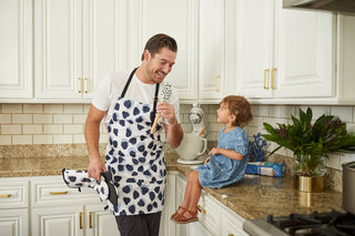 A man in a patterned apron, holding an SDS Spatulas heat-resistant silicone spatula, chats with a blue-clad toddler on the kitchen counter amidst white cabinets, a plant, and a pasta box. The toddler gazes at him curiously.