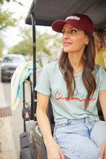 A woman in a Charleston Dad Cap, Maroon, with an adjustable backstrap and a light green T-shirt featuring colorful text sits on a golf cart. She smiles while looking to the side, with trees and a vehicle blurred in the background.