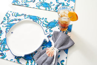 A fish-shaped white plate rests on a Reversible Crab Craze Placemat, beside a glass of iced tea garnished with an orange slice. A seashell-ringed napkin is nearby, creating a coastal flair and charming nautical theme.