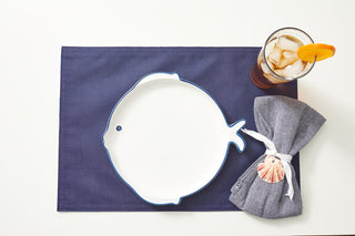 A fish-shaped white plate with a blue rim is on a dark blue Reversible Crab Craze Placemat. To the right, theres iced tea with orange slice and a nautical-themed blue napkin wrapped in a shell decoration, enhancing the coastal look of this light surface setting.