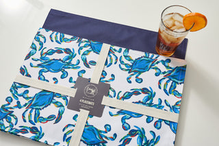 The Reversible Crab Craze Placemat set of four, tied with cream ribbon, features a coastal blue crab design and adds seaside charm next to an iced tea garnished with an orange slice on a white surface.