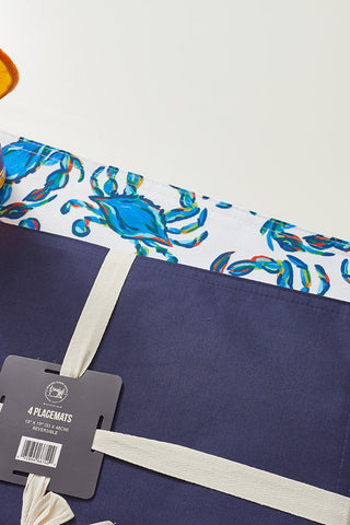 A glass of iced tea with an orange slice sits on two placemats: one is a solid navy with a ribbon and tag, and the other from the Reversible Crab Craze Placemat set showcases a charming blue crab pattern. They rest elegantly on a pristine white surface.