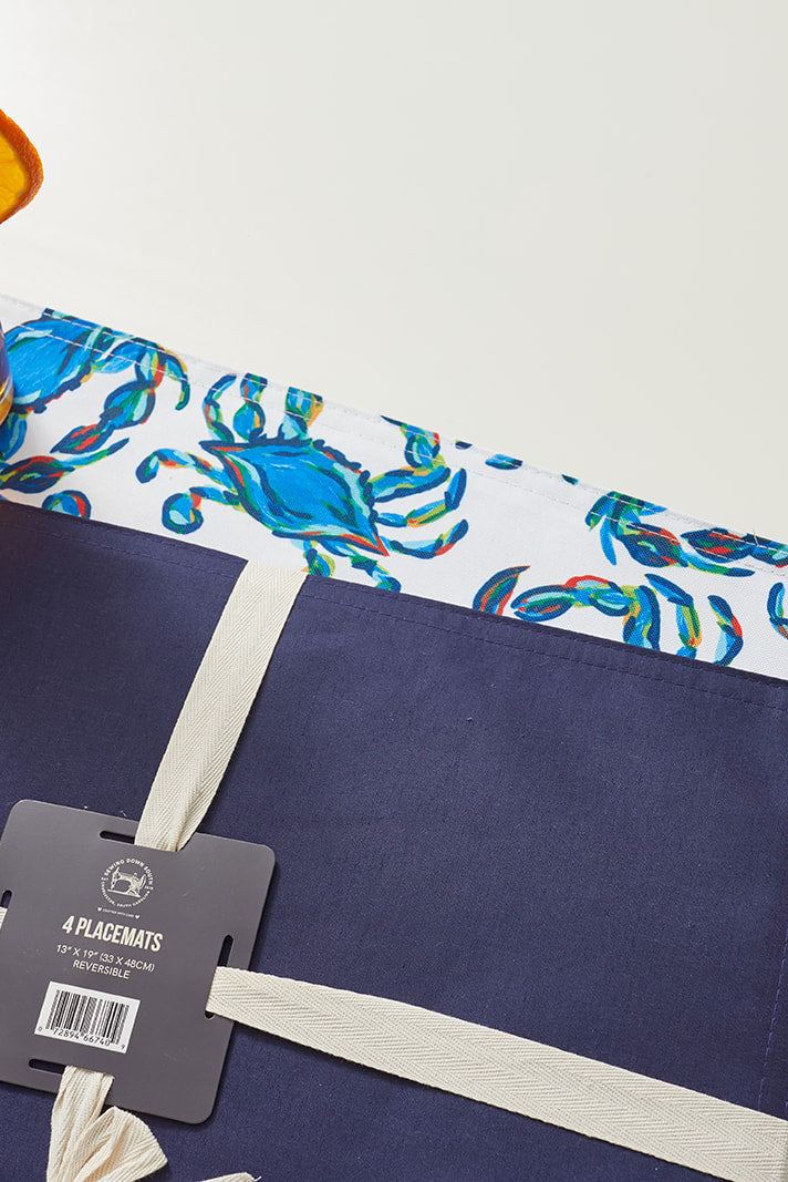 A glass of iced tea with an orange slice sits on two placemats: one is a solid navy with a ribbon and tag, and the other from the Reversible Crab Craze Placemat set showcases a charming blue crab pattern. They rest elegantly on a pristine white surface.