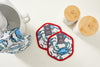 Two Crab Craze hexagonal pot holders with blue crab designs, red borders, and silicone grips lie on a white surface, featuring labels. Nearby are a ceramic pitcher and two jars with bamboo lids, while a Crab Craze-patterned cloth peeks in from the left.