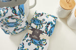 The Crab Craze Kitchen Towels, 2 pack, showcases a vibrant crab design alongside a chic white jar and textured container with a wooden lid. A pot glimpse adds charm, making it perfect for Coastal Kitchen Decor.