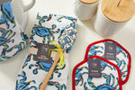The Crab Craze collection includes a blue crab design on kitchen items like pot holders with red trim and silicone grip, tea towel sets, a wooden spoon, and ceramic containers with bamboo lids, all elegantly displayed on a white surface.