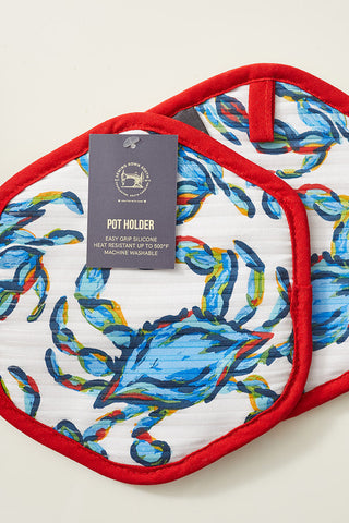 The Crab Craze pot holders, featuring two hexagonal pieces with a vibrant blue crab design and red trim, are perfect for enthusiasts. One has a Pot Holder tag with care instructions. With silicone grip and heat resistance, they are both functional and stylish against a plain light surface.