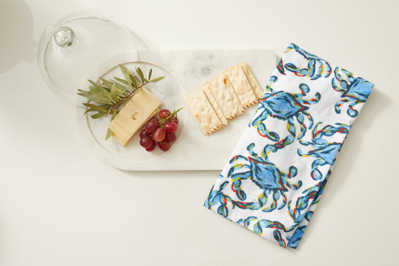 A marble serving board elegantly displays cheese, grapes, and green leaves. A rectangular cracker complements the scene, while a neatly folded Crab Craze Kitchen Towel from a 2-pack set with blue and green designs adds a touch of color beside the board.