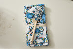 The Crab Craze Kitchen Towels, a 2-pack with a vibrant blue, red, and yellow crab pattern, are neatly folded and tied with a beige ribbon. Tucked under the ribbon is a wooden-handled spatula embodying Coastal Kitchen Decors charm, elegantly placed on a light surface.
