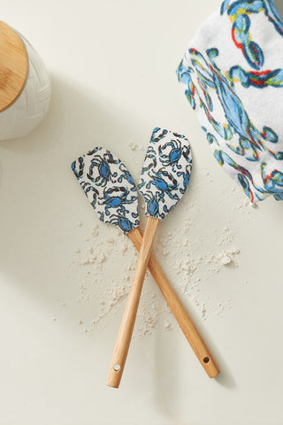 On a flour-dusted countertop, two crossed SDS Spatulas with wooden handles and blue crabs rest among nonstick cookware. Nearby are jars with wooden lids, a patterned towel, and mixer parts—ideal for any culinary adventure!.