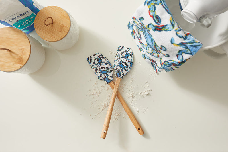On a flour-dusted countertop, two crossed SDS Spatulas with wooden handles and blue crabs rest among nonstick cookware. Nearby are jars with wooden lids, a patterned towel, and mixer parts—ideal for any culinary adventure!.