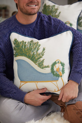 A man in a blue sweater holds the Sewing Down South Sleigh Pillow, which features a blue sleigh carrying a Christmas tree and a yellow dog in a scarf.