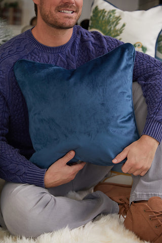 A man in a purple cable-knit sweater and light gray pants sits on a fuzzy rug, holding the Sleigh Pillow with an exclusive Sewing Down South pattern in Parisian blue velvet on his lap. Part of his brown boots are visible.