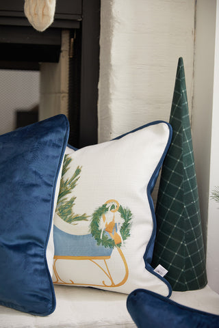 A Sleigh Pillow from Sewing Down South sits beside a blue velvet pillow cover and a green plaid cone-shaped ornament, all arranged in front of a white brick wall.