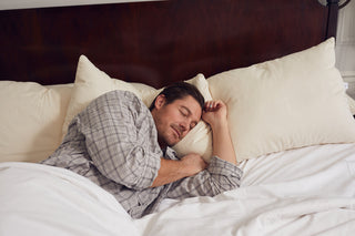 A man sleeps peacefully on his side in a bed with white sheets and large pillows, resting his head on the SDS Organic Sleeping Pillow, King, which features hypoallergenic fill for comfort and relaxation.
