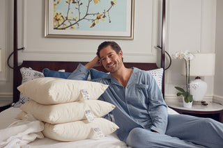 A man in blue pajamas sits on a neatly made bed, smiling and leaning on a stack of SDS Organic Sleeping Pillow, Queen. The GOTS Certified organic cotton pillows have visible tags; a painting and white orchid decorate the room.