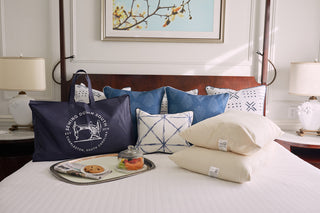 A neatly made bed with blue and white decorative pillows, two SDS Organic Sleeping Pillow, Queen with hypoallergenic fill, a navy tote bag, and a silver tray holding cookies, fruit, and a drink. Lamps and artwork are in the background.
