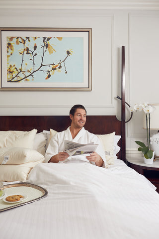 A man in a white robe sits up in bed smiling while reading a newspaper. The bed features GOTS Certified Organic Cotton linens and the SDS Organic Sleeping Pillow, King. A breakfast tray is on the bed, with a floral painting hanging behind him.