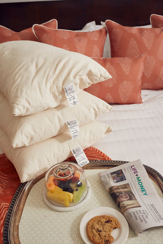 Three SDS Organic Sleeping Pillow, King pillows with visible tags are stacked on a bed topped with coral-patterned cushions. In front, a tray holds a covered fruit bowl, a cookie on a plate, and a LIFE+MONEY newspaper section.