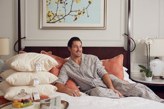 A man in plaid pajamas sits smiling on a neatly made bed with orange pillows and a stack of SDS Organic Sleeping Pillow, King (GOTS Certified Organic Cotton). A bedside table holds flowers, fruit, and a lamp beneath framed artwork.