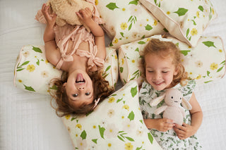 Two girls are smiling joyfully on a bed with Kids Collection: Pink Floral Pillow. One, with curly hair, cuddles a beige teddy bear, while the other, with wavy hair, holds a bunny toy.