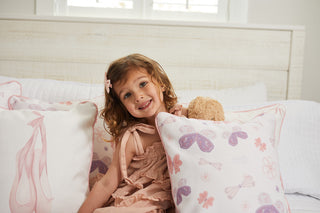 A young girl with curly hair and a pink bow from the Kids Collection beams while sitting on a bed in natural light. Shes wearing a light pink dress, holding a teddy bear, and surrounded by Bow & Ballet pillows from Sewing Down South®.