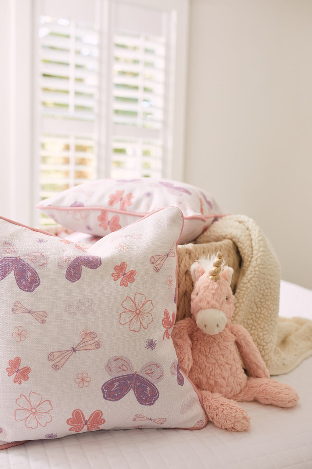A cozy bedroom scene showcases a cute pink unicorn plush toy nestled beside two Butterfly Pillows from the Kids Collection, ideal for nursery decor. A beige knitted blanket drapes behind them, and white shutters illuminate with soft light. Perfect for any kids collection!.
