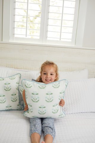 A light brown-haired child sits on a white bed clutching a Victorian Floral in Blue Pillow from the Kids Collection, smiling at the camera. The bed is adorned with patterned pillows that match, and soft daylight filters through white shutters.