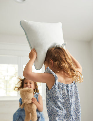 Two young girls joyfully have a pillow fight in a bright room, wearing charming dresses. One holds a Kids Collection: Victorian Floral in Blue Pillow above her head, while the other clutches a teddy bear and grins widely.