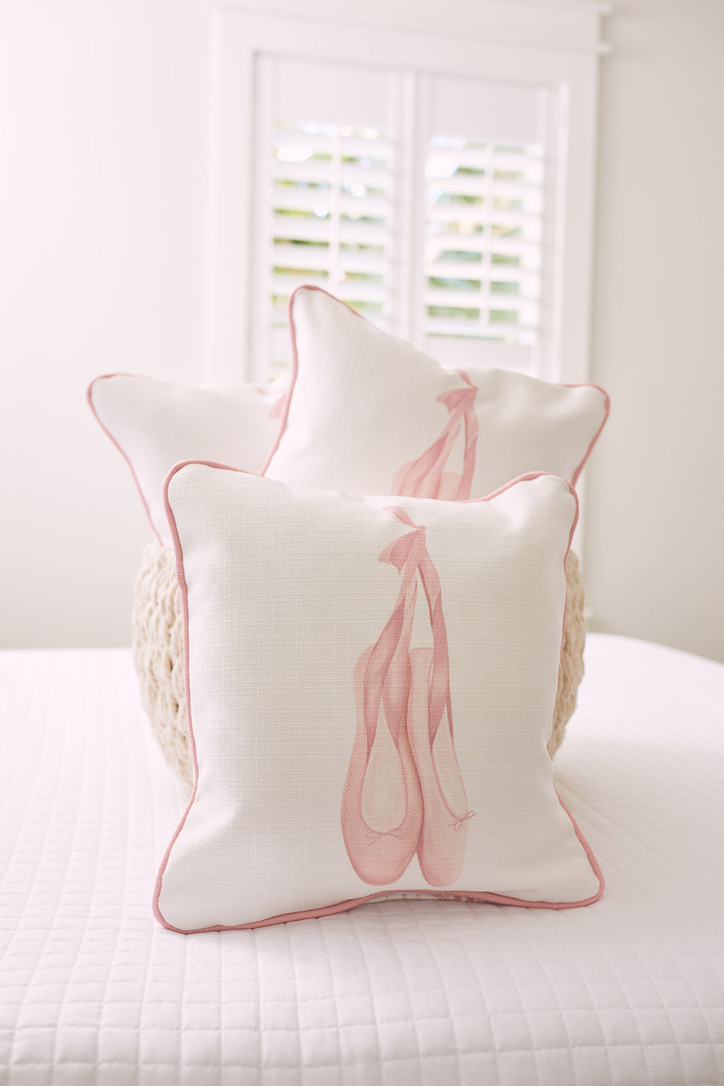 Three white pillows from the Kids Collection, featuring pink piping and ballet shoe illustrations, are stacked neatly on a white quilted bed. The Bow & Ballet pillow by Sewing Down South® catches soft light streaming in through the window with its distinct pattern and design.