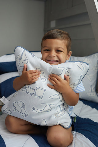 A young child with short brown hair sits on a bed, smiling widely and hugging a Kids Collection: Transportation Pillow adorned with blue and white car, train, and boat patterns that perfectly complement the striped bedding.