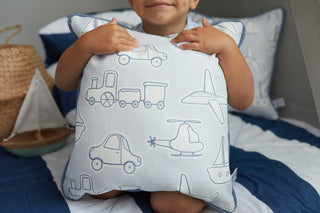 A child holds a Kids Collection: Transportation Pillow featuring blue outlines of cars, trains, airplanes, and helicopters. The scene is completed with blue and white striped bedding, a straw hat, and a model sailboat from Sewing Down South®.