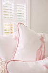 A close-up showcases a white cushion with light pink piping and abstract pink patterns from the Kids Collection: Bow & Ballet Pillow. The backdrop of softly focused white shutters creates a serene ambiance.