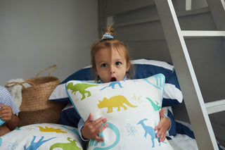 A young child with a surprised expression holds the Kids Collection: Dinos Pillow from Sewing Down South®, featuring colorful dinosaurs, standing beside a bunk bed and woven basket in their playful, cozy room.