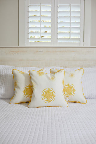 Three pillows from the Kids Collection: Sun Pillow, featuring yellow sunburst patterns, rest on a white bed. They create a charming scene in the softly lit room, arranged against a window with shutters.