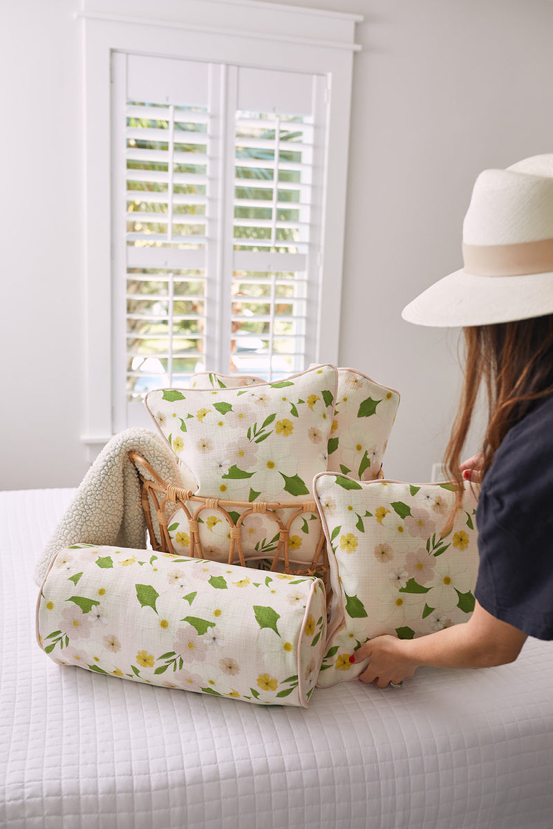 A woman in a hat places a Pink Floral Pillow from the Kids Collection and its matching blanket into a wicker basket on the bed. The rooms white walls and shuttered windows fill with natural light, enhancing the bright and airy decor.