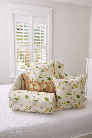 A neatly arranged pile of decorative pillows and cushions, including the Kids Collection: Pink Floral Pillow with green leaves and yellow flowers, sits atop a white bedspread. Visible in the background through a window with white shutters is greenery outside.