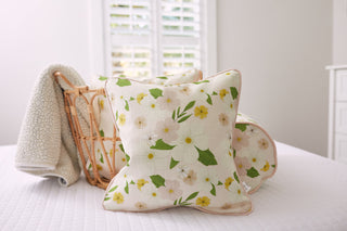 The Kids Collection: Pink Floral Pillows, adorned with white and yellow flowers, rest in a basket on a white quilted bedspread. A light-colored throw hangs over the baskets side as soft natural light streams through shuttered windows.