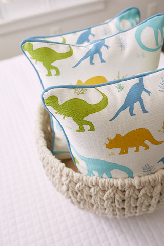 Two Kids Collection: Dinos Pillows in a textured woven basket display vibrant green, blue, and orange dinosaurs on a light background, creating a bright and minimalistic setting.