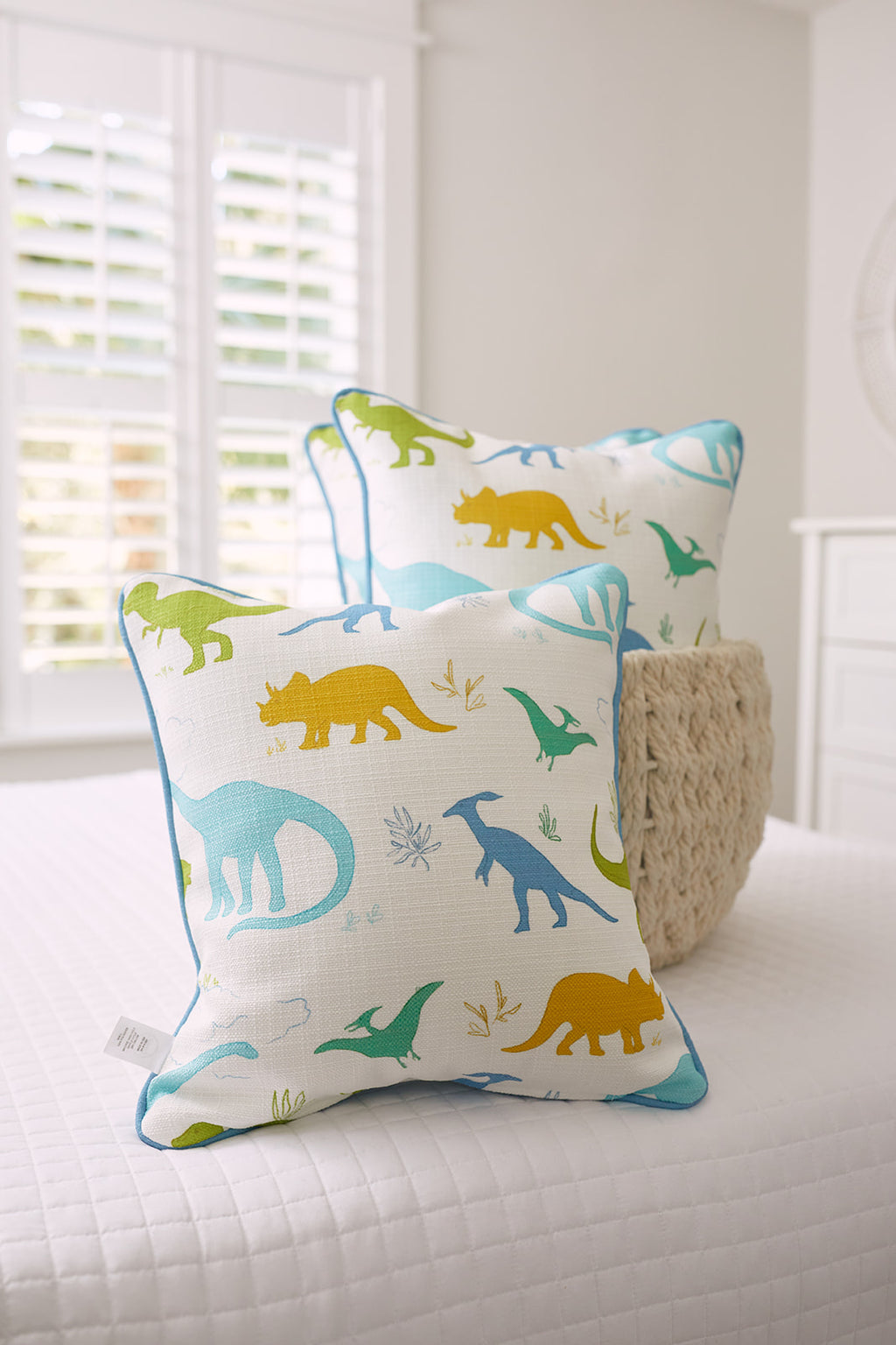 Two Kids Collection: Dinos Pillows from Sewing Down South® adorn a white bed, featuring green, blue, and yellow dinosaur silhouettes. A woven basket sits nearby, with a window framed by white shutters adding to the playful scene.